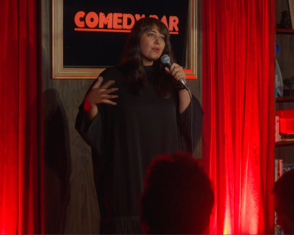 Geetika Khurana on stage at a comedy club, mid-joke with a microphone in one hand, red curtains and a neon Comedy Bar sign behind her.